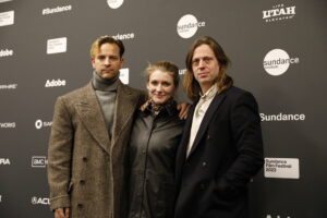 A blonde man in a grey turtle neck and a brown coat, a blonde woman in a black coat, and a man with long brown hair in a blakc coat, stand in front of a step and repeat at the 2023 Sundance Film Festival