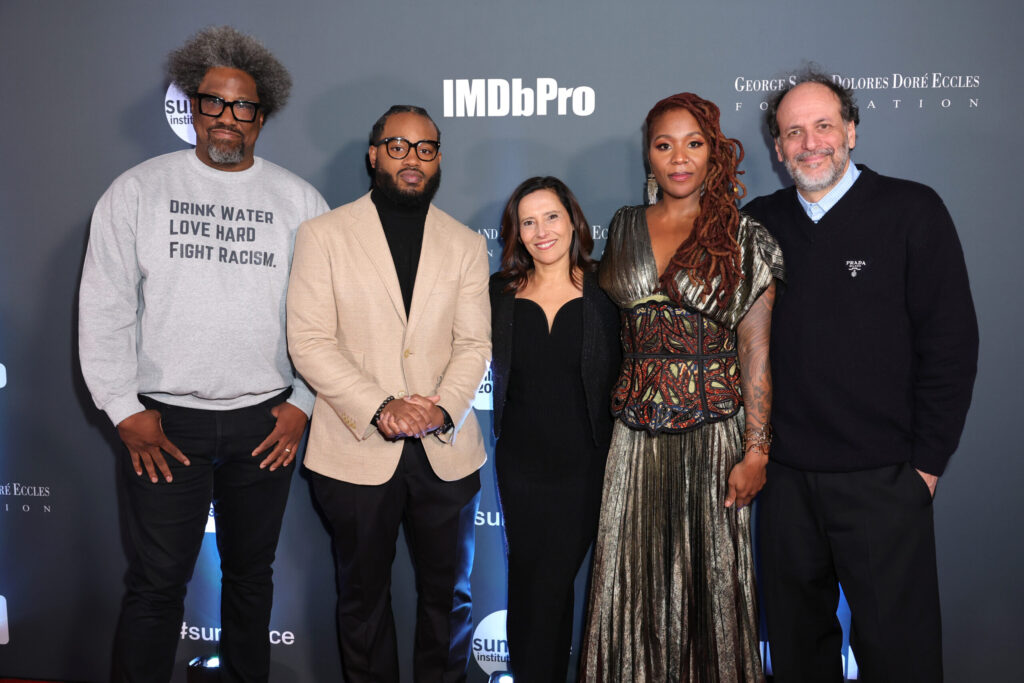 From left to rigth: A black man with glasses and a grey sweatshirt, a Black man with a beard and a cream jacket, a shorter woman with brown hair and a black dress, a Black woman with long hair and a gold dress, and a tall italian man with grey hair and a black jacket are standing in front of a step-and-repeat
