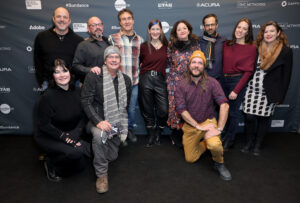 The production team for Justice, ten members total, are standing in front of the Sundance Film Festival black step-and-repeat two men in beanies and a woman in a black coat are kneeling in front.