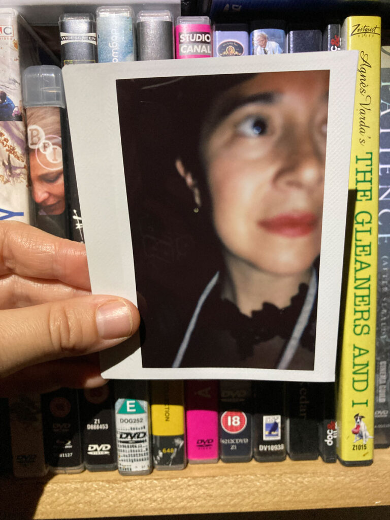 white hand holding a polaroid of a white woman's face up in front of a shelf of blu rays