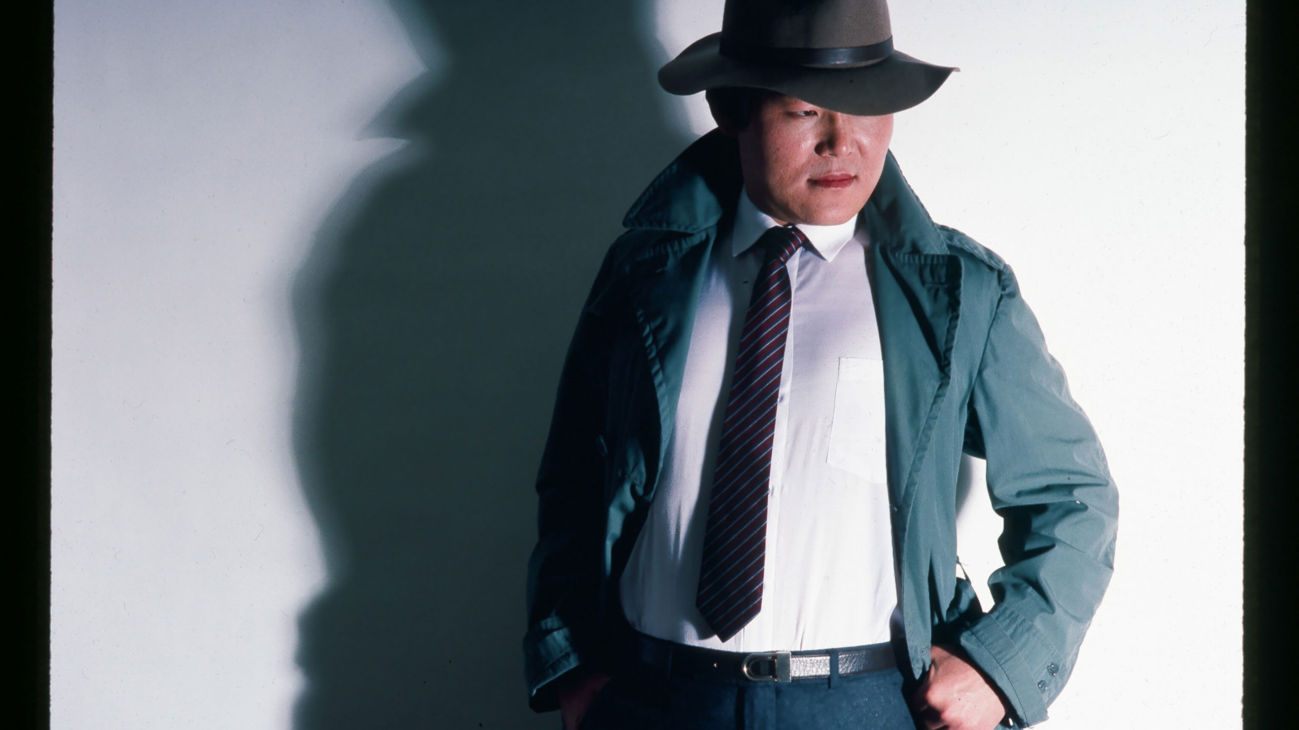 asian man in a jacket and tie with a fedora on looking to the right against a white wall