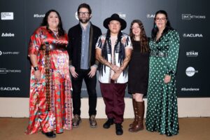 A woman with long black hair in a red dress, a dar-haired man with glasses abd a black jacket, a woman in long braids and a a white and blue shirt and red pants, a woman with long brown hair and a black dress with brown boots, and a woman in a lok green polka dot dress, stand in front of a step and repeat at the 2023 Sundance Film Festival.