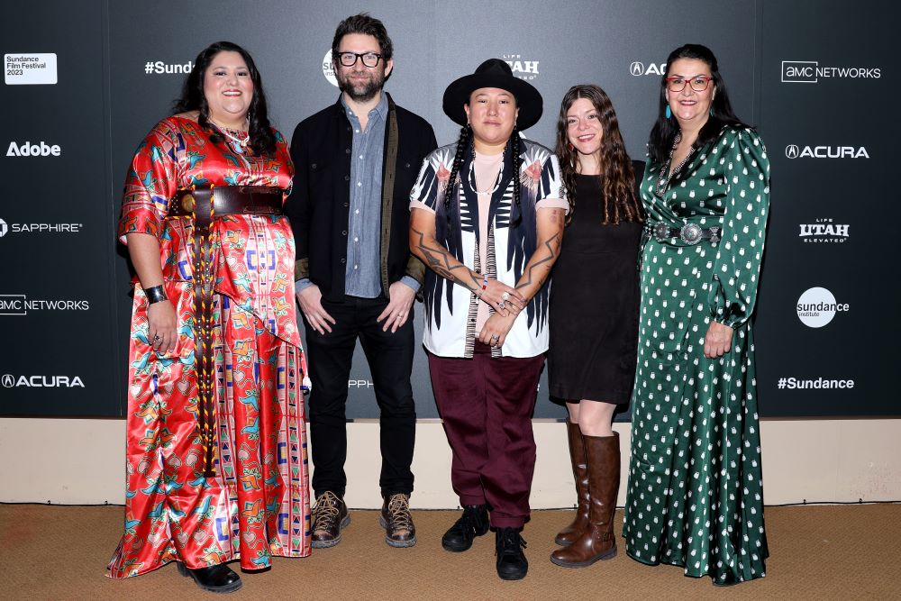 A woman with long black hair in a red dress, a dar-haired man with glasses abd a black jacket, a woman in long braids and a a white and blue shirt and red pants, a woman with long brown hair and a black dress with brown boots, and a woman in a lok green polka dot dress, stand in front of a step and repeat at the 2023 Sundance Film Festival.