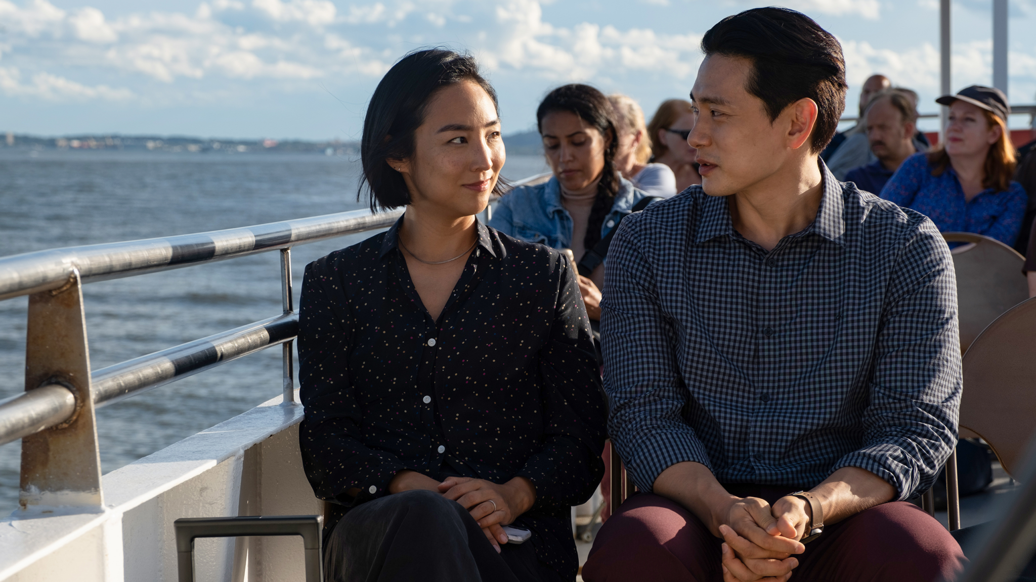 Young Asian woman and man sit side-by-side looking at each other, on a boat also transporting other people.