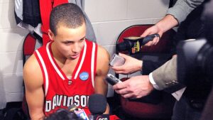 A young, bald basketball player in a red jersey speaks to reporters