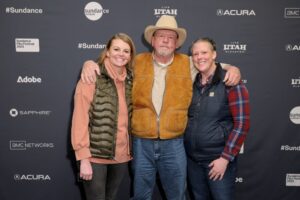 A blonde woman in a peach-colored long-sleeved shirt with a gold vest, a man with a yellow jacket vest in a cowboy hat, and a woman in a black vest over a red plaid long-sleeved shirt with her hair tied back, stand in front of a step and repeat at the 2023 Sundance Film Festival.
