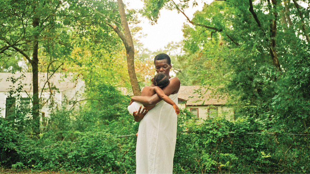 A black woman wearing all white is holding a baby close to her in a green forest