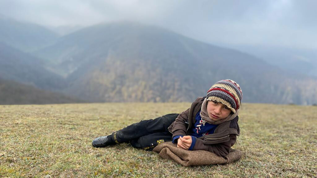 A young boy with long pants, a coat, and a colorful cap lays on a folded blanket on the grass with a mountain in the background