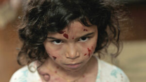 A a close-up of a young girl in a white shirt with black hair and blood on her face as she stares into the camera.