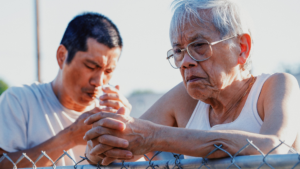 a young Asian man in a white tshirt smokes a cigarette next to an older Asian man with glasses and grey hair in a white tank top