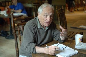 Older man with gray hair, sitting at a table, wearing a gray sweater and holding his spectacles, gestures as if speaking to someone off-camera.