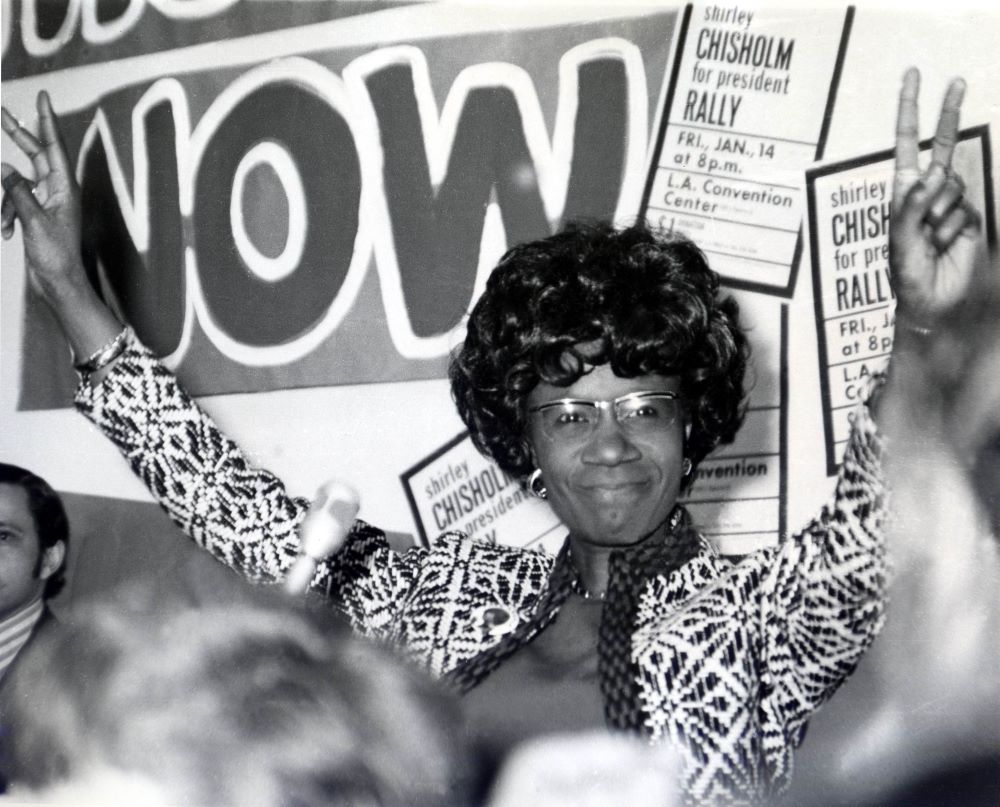 Black woman wearing eyeglasses and a print dressy jacket smiles, with both arms raised and fingers making the victory sign, in front of campaign posters.