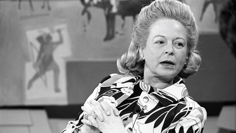 White woman with bouffant hair and print dress, looking off-camera as if being questioned or interviewed