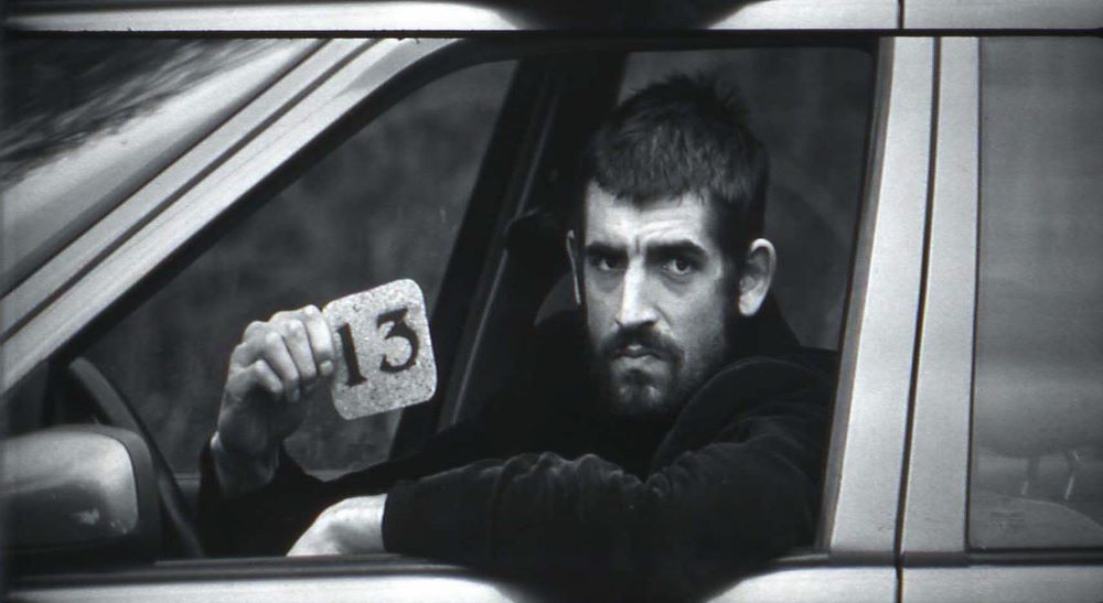 A dark-haired, bearded man stares somberly out the driver's side window of a car, holding a card with the number 13.