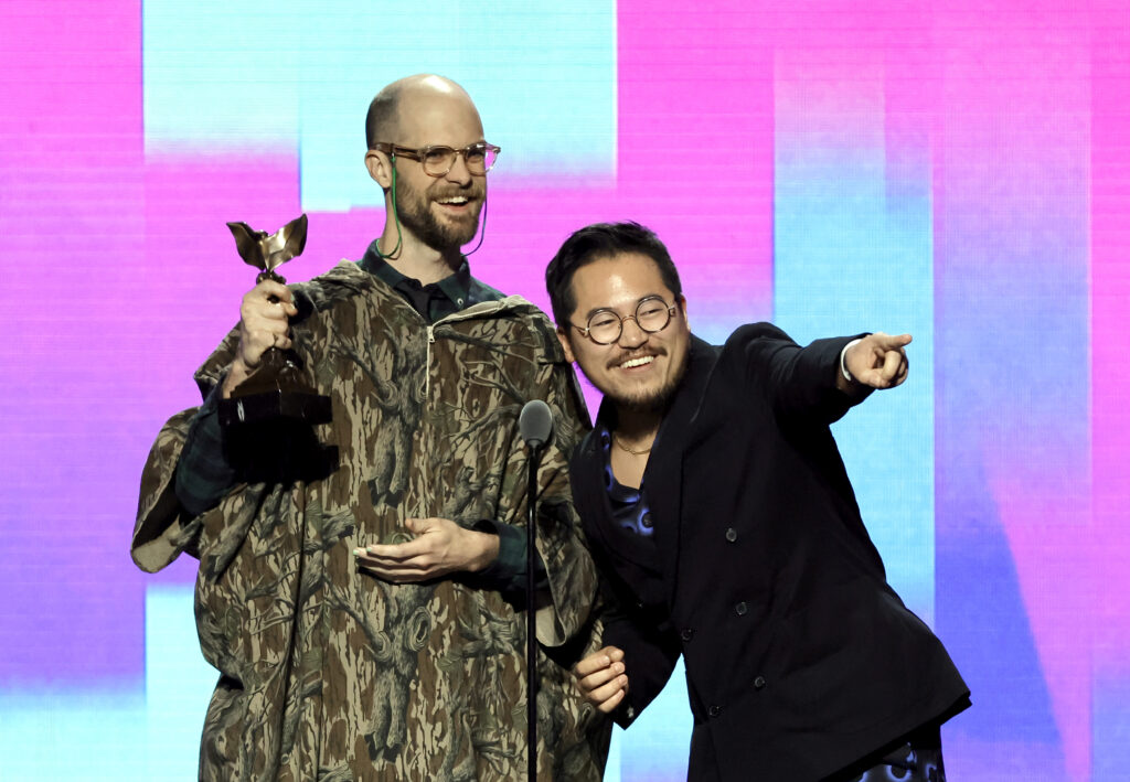A caucasian bald man holds a Spirit Award next to an Asian man with black hair reaching to the audience