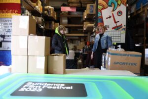 Two women stand in a warehouse setting, with a Sundance Film Festival 2023 poster in the foreground.