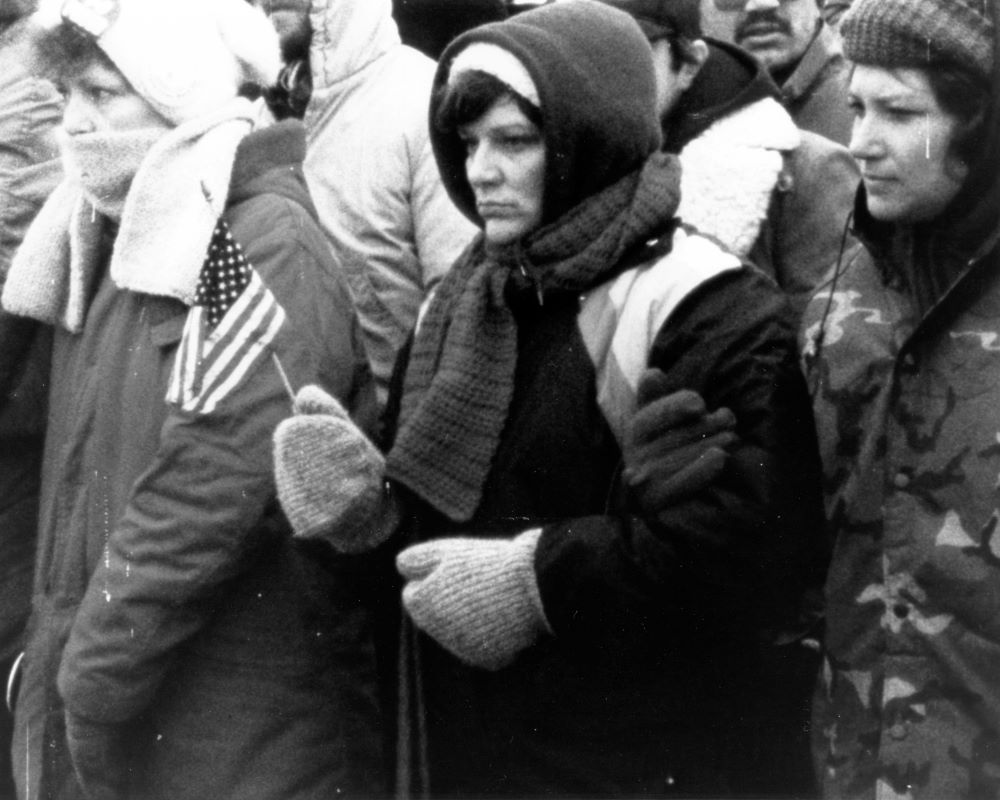 A woman dressed in heavy coat and gloves holds a small American flag while standing in a group of similarly dressed people