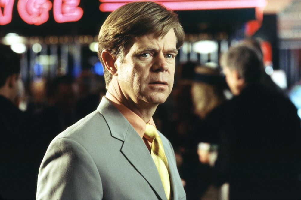 A brown-haired man in gray suit, gold tie, and pale orange shirt is shown in a head-and-shoulders photo, standing in front of what appears to be a night scene with pink neon lights.