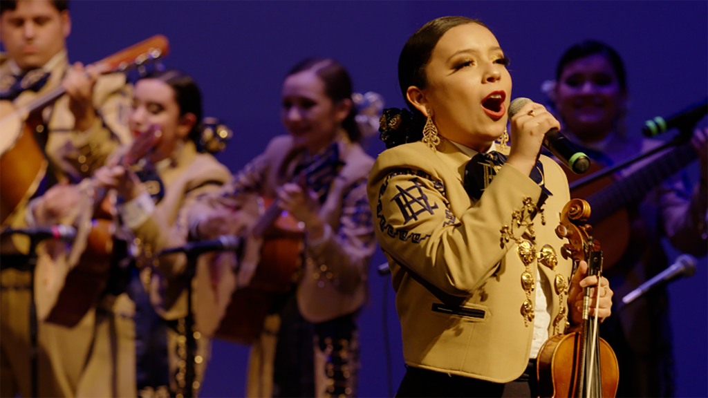 A Mariachi singer dressed in gold with her hair tied back is singing into the mic.