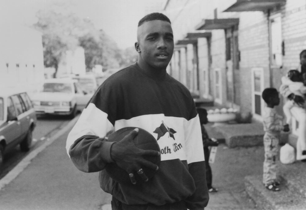 Young Black man, cradling a basketball in one big hand, stands in a city neighborhood