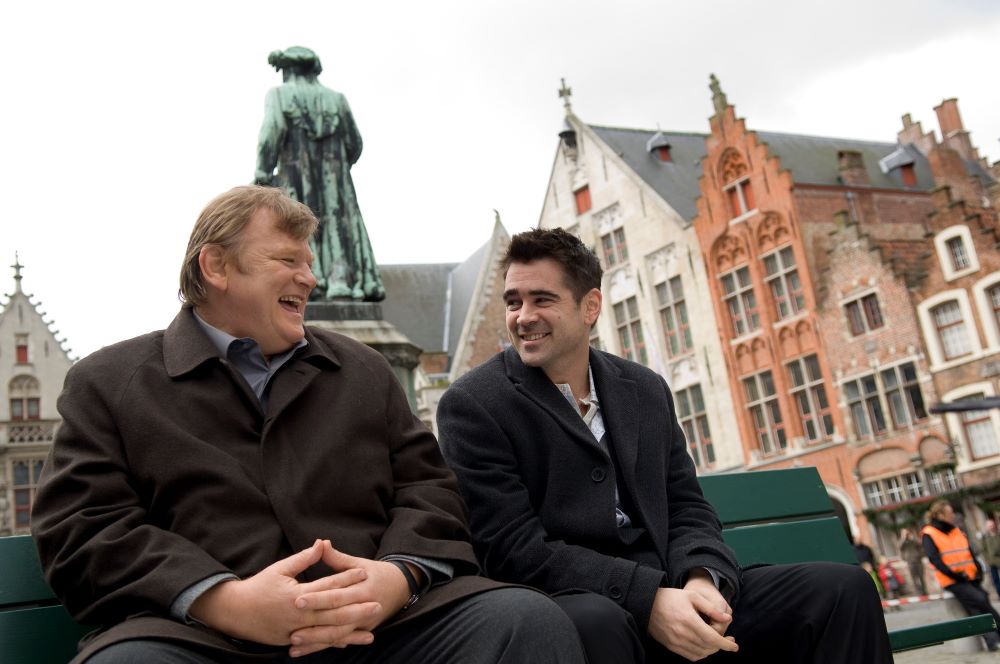Two men, one a larger man with brown hair, the other with darker hair, sit on a bench, smiling and laughing. The setting appears to be a European village