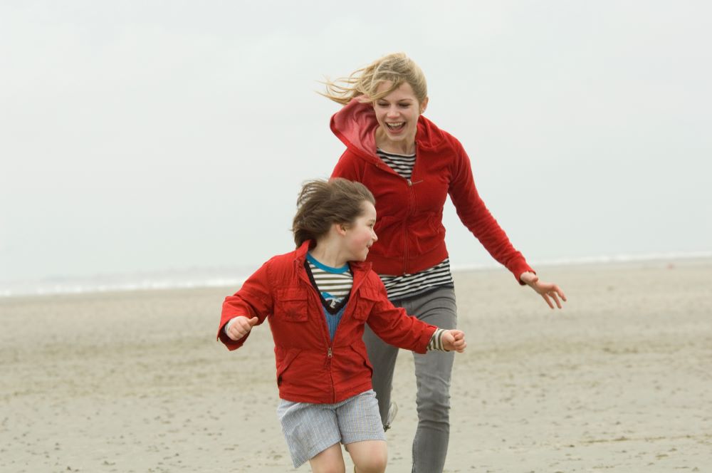 Blond woman in red hoodie chases a brown-haired boy in shorts and red hoodie along a beach. Both are laughing.