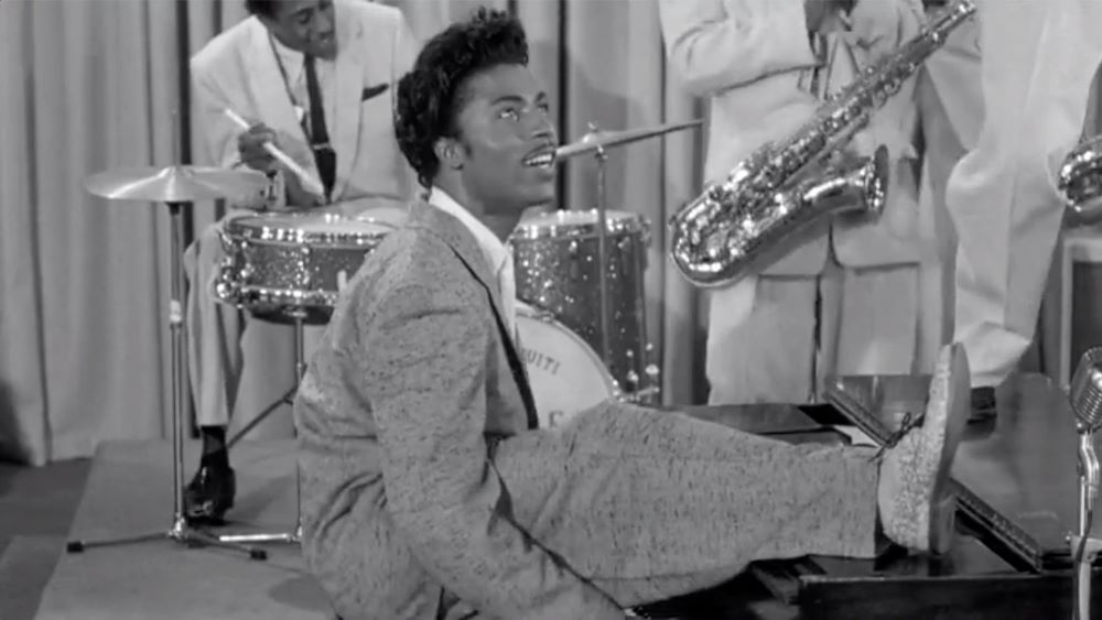 Man in a light tweed-looking suit at a piano, one leg thrown up over the top of the instrument, in front of band members, including a drummer and a drum set and a man holding a saxophone