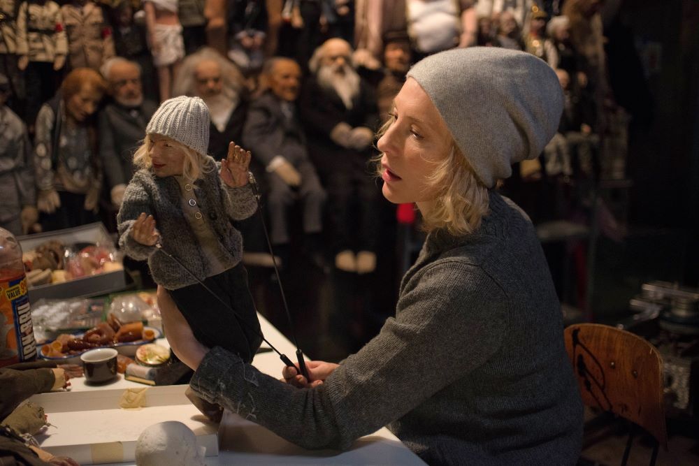 Blond woman in stocking cap examines a puppet that looks like her and is dressed like her. Puppets line the shelves behind her.