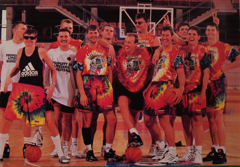 A jubilant basketball team standing on a basketball court for a team picture, flashing victory signs, many wearing or holding colorful tie-dye basketball shorts