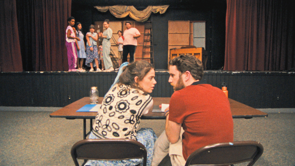 A young woman with brown hair talks to a young man with brown hair and an orange shirt. A stage is in the background with theatre campgoers looking on