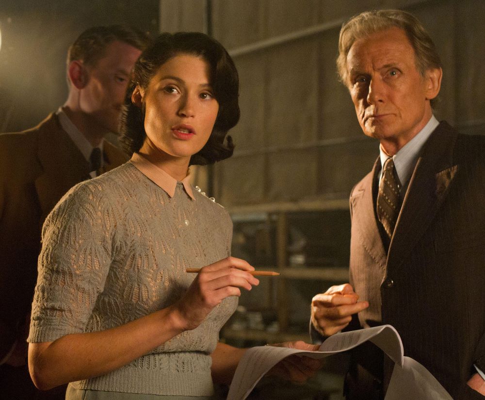 Woman with dark hair, holding a pencil and some stapled pages, stands beside an older, gray-haired man in a suit. Both are looking at something in front of them