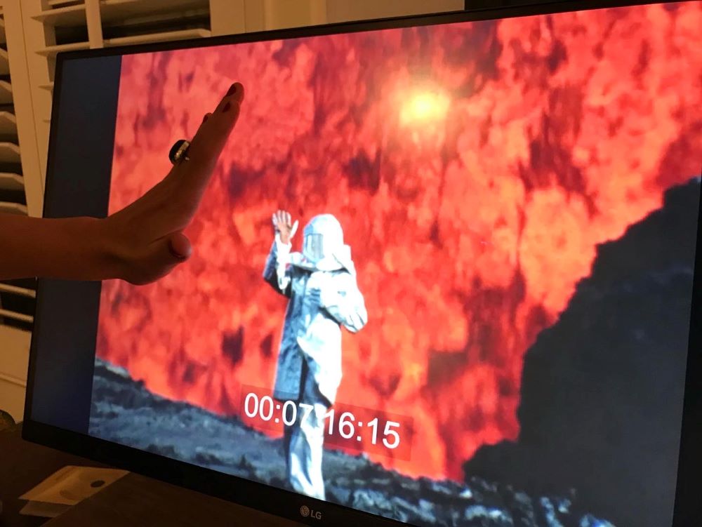 Person in protective suit with fiery background appears on computer screen. The person is waving, and someone in the room with the computer returns the wave; only the hand is in the frame.