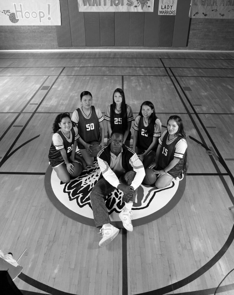 Young Native American girls sit in an arc on the center logo of a basketball court, with a Black man inside their arc