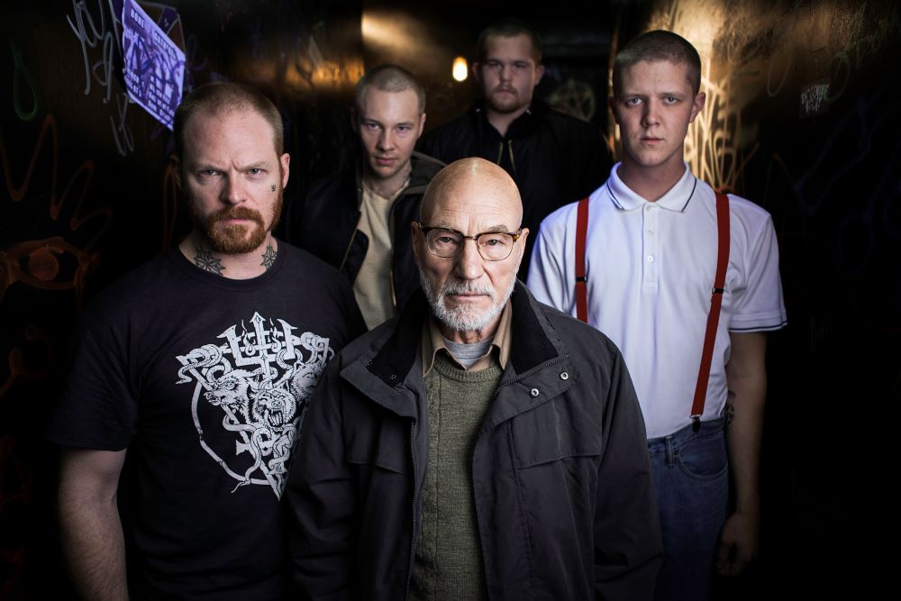 Four men with closely shorn hair, one with tattoos on his face and neck, standing in a group fronted by a bald, gray-bearded older man wearing eyeglasses. Background is dark, perhaps a nightclub.