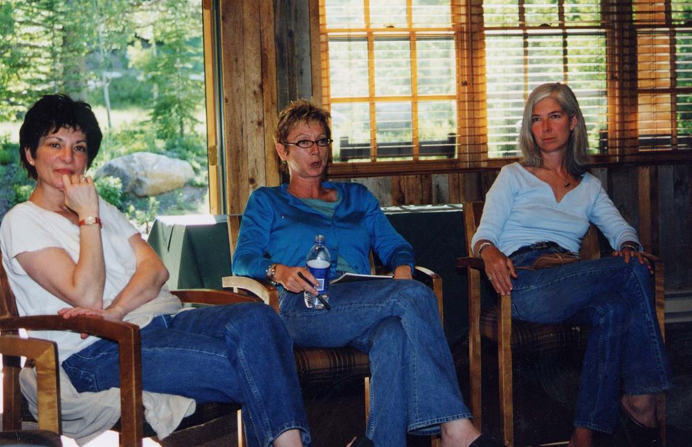 Three women sit in an arc of chairs in a room with evergreen trees and a path outside the window. All are dressed casually in jeans.