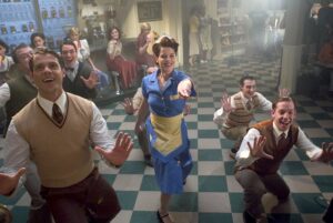 A woman in a blue dress dances in the middle of five men on a checkered floor.