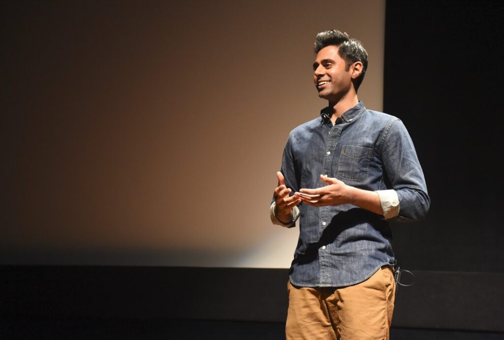 Comedian Hasan Minhaj stands in front of a screen smiling wearing a blue shirt and tan pants