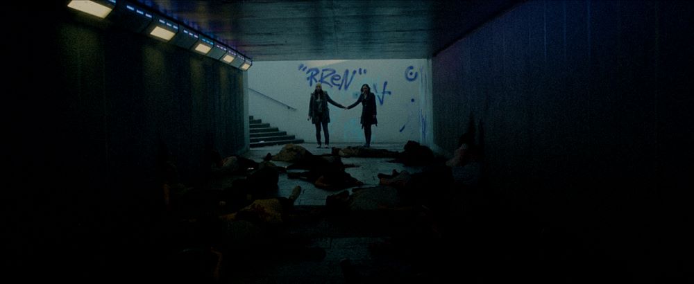 Two women, holding hands, stand at the bottom of a stairs leading to what appears to be a concrete tunnel. In the foreground are what appears to be bodies strewn in front of them
