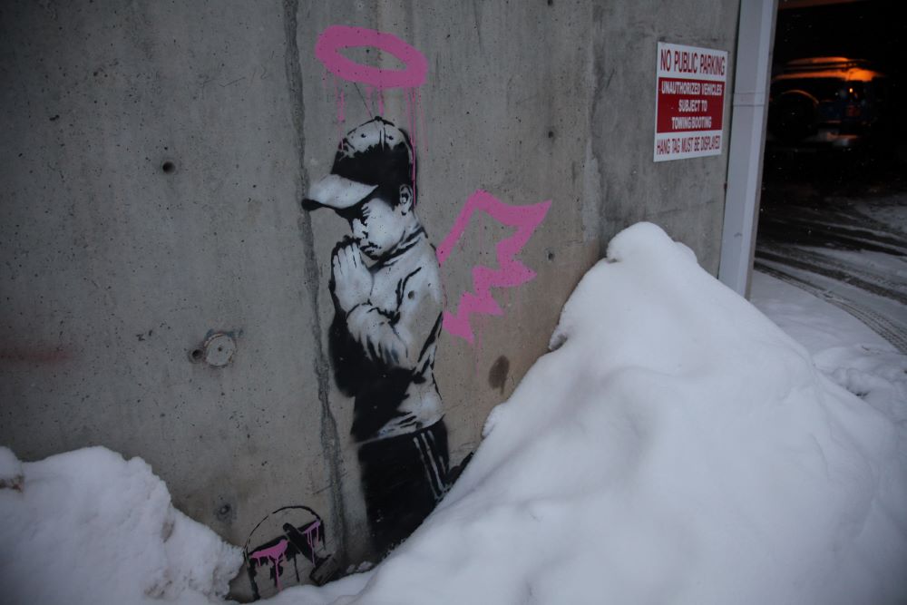 On a gray exterior wall leading to a parking garage is a painting of a boy wearing a ball cap, on his knees, hands folded as if in prayer. He has a pink halo and pink angel wings, and a bucket of pink paint in front of him