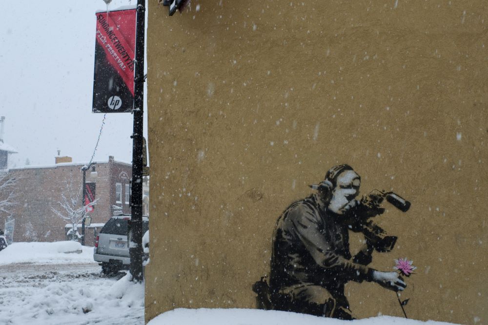 With snow coming down on a city street, a yellowish building wall displays a painting of a cameraman pointing his lens at an uprooted pink flower. To the left, on a street pole, a red and black banner announces the Sundance Film Festival.