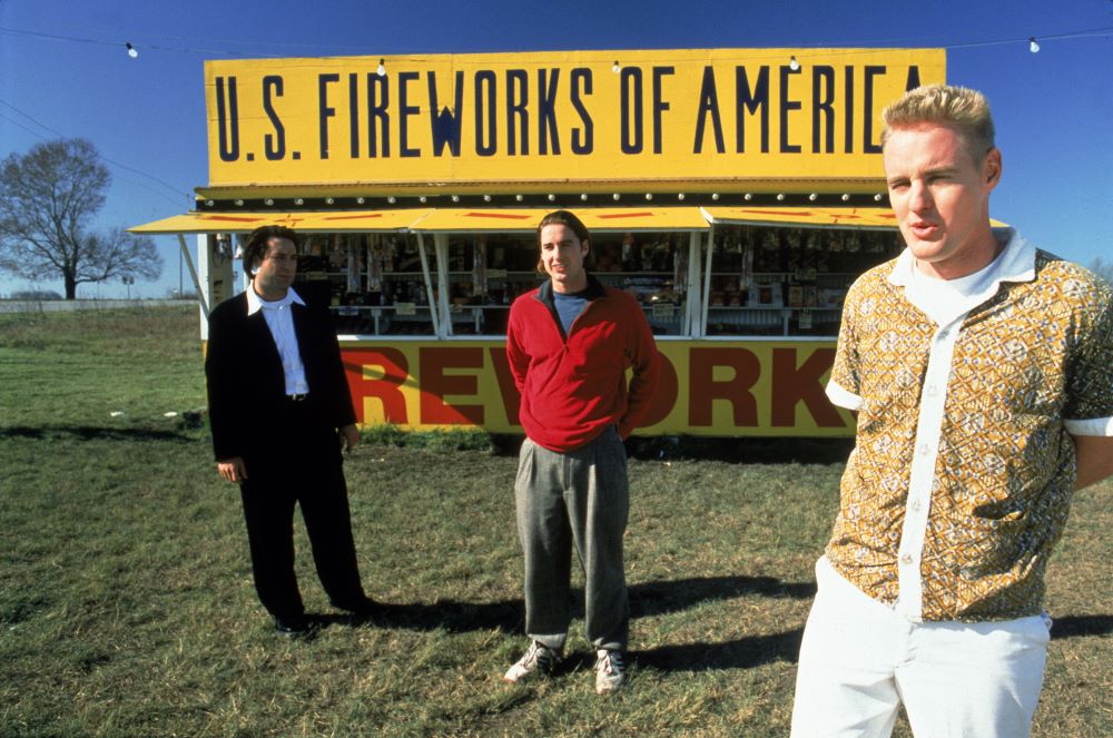 Three men stand casually, spaced apart, in front of a fireworks stand