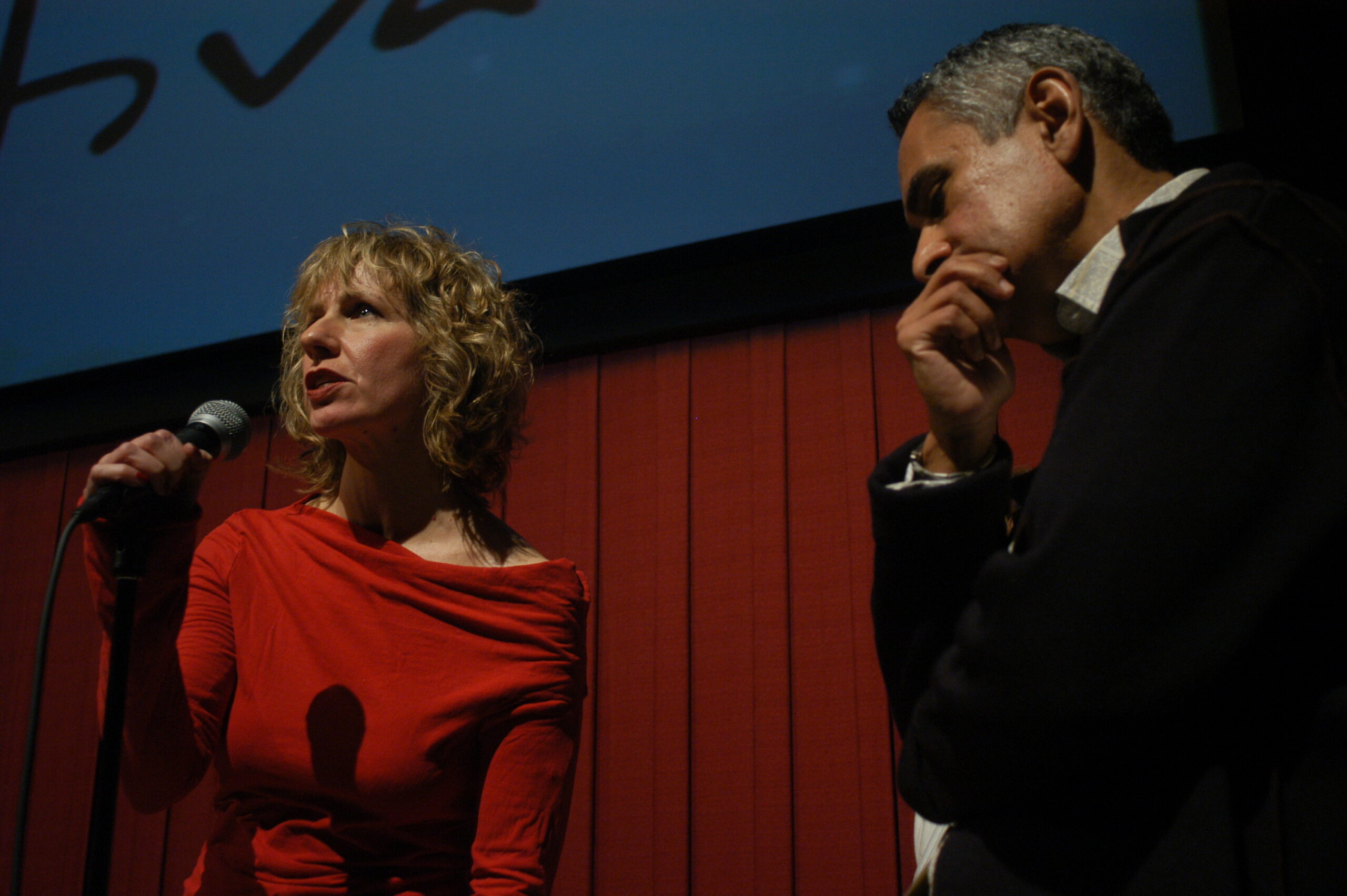 A woman in red with short blonde hair speaks into a mic in front of a large screen. Next to her is a Hispanic man with grey/black hair wearing a suit.