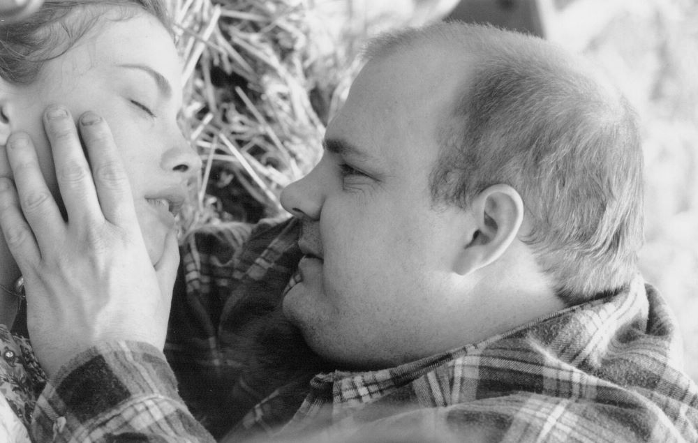 Younger man with short light brown hair who appears to be overweight caresses with his left hand the cheek of a young woman whose eyes are closed