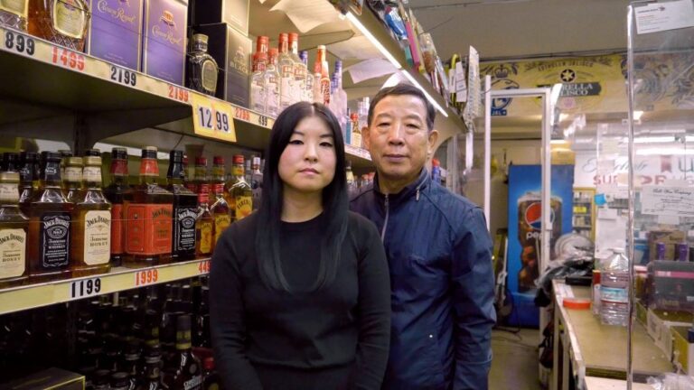Young Asian woman and older Asian man stand beside shelves holding liquor bottles in a store.