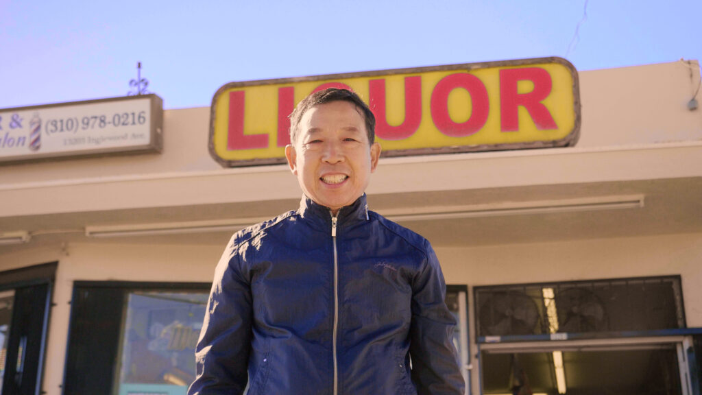 Asian man in dark blue jacket stands, smiling broadly, in front of a liquor store