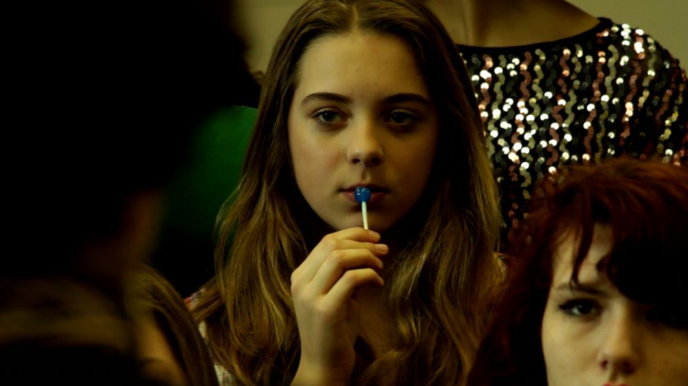At the center of this image is a young woman with long, brown hair, holding a blue sucker to her lips