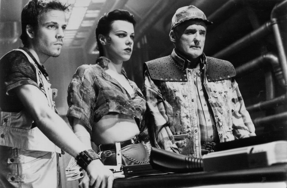 A young man, a woman, and an older man in unusual attire stand shoulder-to-shoulder, staring at something in the right foreground