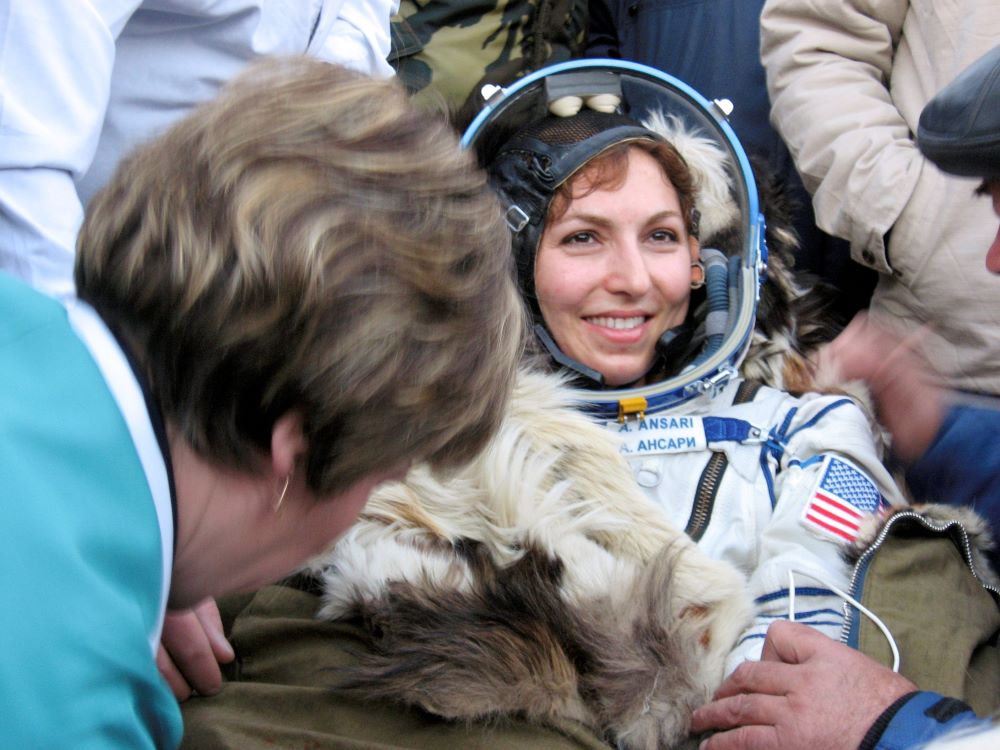 Woman in astronaut gear smiles, surrounded by other people and a furry animal