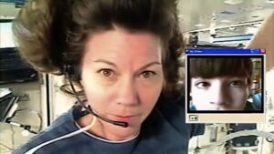 Woman appears to be floating in zero gravity in a spaceship. She wears a headset and appears to be talking with a young boy, who appears in an inset on the photo.