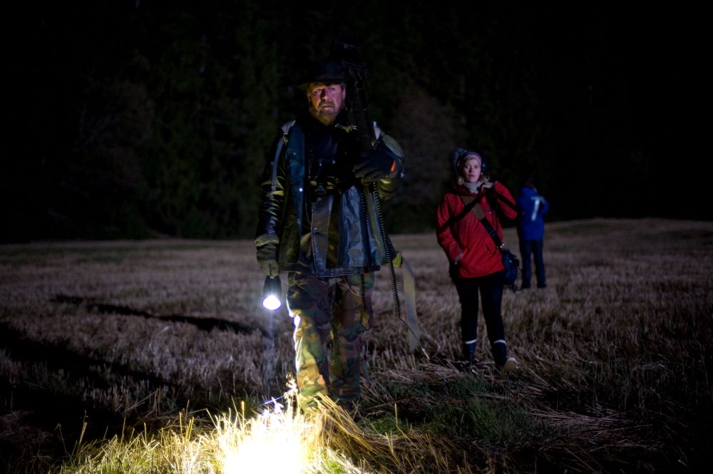 Nighttime shot in a field with a bearded man in camo and holding a flashlight, a woman in a red parka following behind, and a person in a blue parka with back to the camera